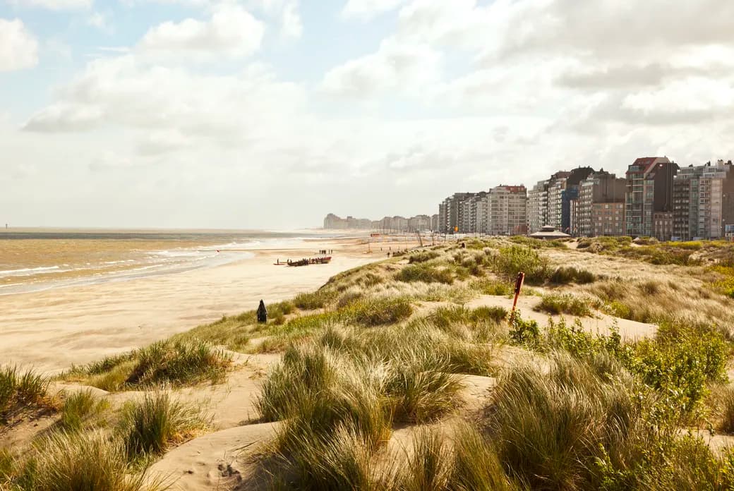 Sandy beach and wind-swept dunes in foreground, small rescue boat and people near shoreline, beachfront apartment buildings under a cloudy sky.