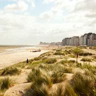 Sandy beach and wind-swept dunes in foreground, small rescue boat and people near shoreline, beachfront apartment buildings under a cloudy sky.