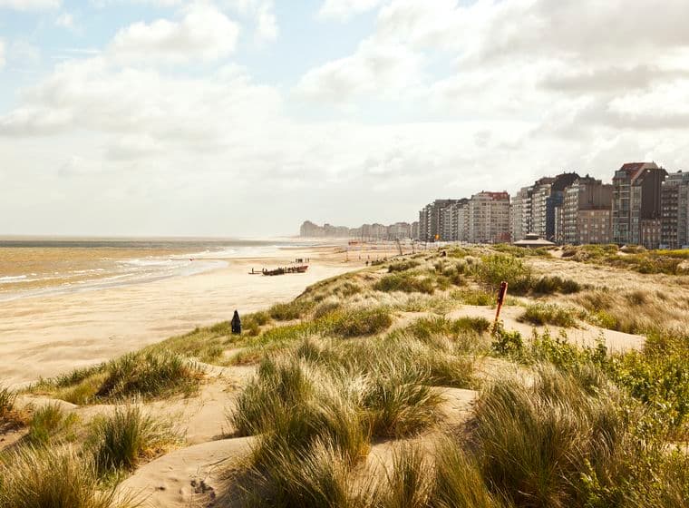 Sandy beach and wind-swept dunes in foreground, small rescue boat and people near shoreline, beachfront apartment buildings under a cloudy sky.