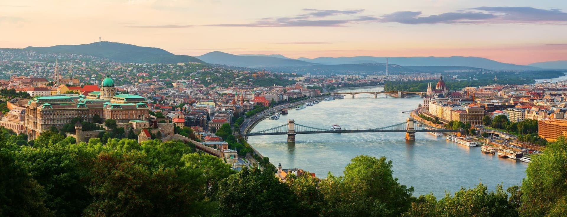 Sunset panoramic view of downtown Budapest: on the left is the Buda Castle building, in the middle is the Danube and the Chain Bridge, and on the right is the Pest side and the Parliament Building with the Buda hills in the background.