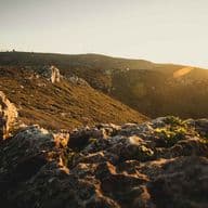 Pôr do sol sobre uma paisagem rochosa com colinas distantes e um céu dourado, criando uma atmosfera quente e serena.