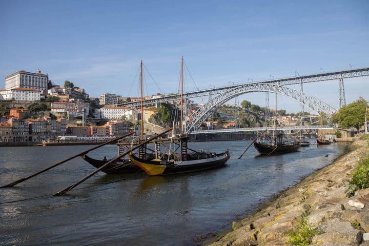 Traditional boats on the Douro River with the Dom Luís I Bridge and hillside buildings in Porto, Portugal, under a clear blue sky.