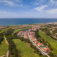 Vista aérea de uma cidade costeira com casas coloridas, campo de golfe verde e o oceano ao fundo sob um céu parcialmente nublado.