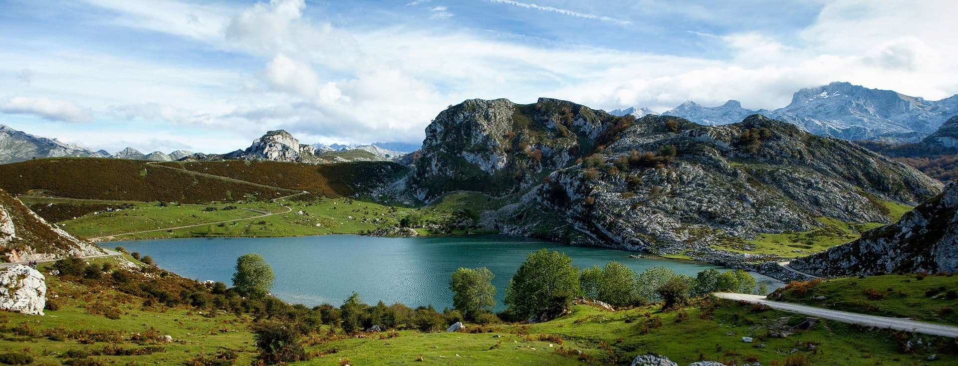 Vista panoràmica d'un llac blau envoltat de turons verds i muntanyes rocoses sota un cel ennuvolat.