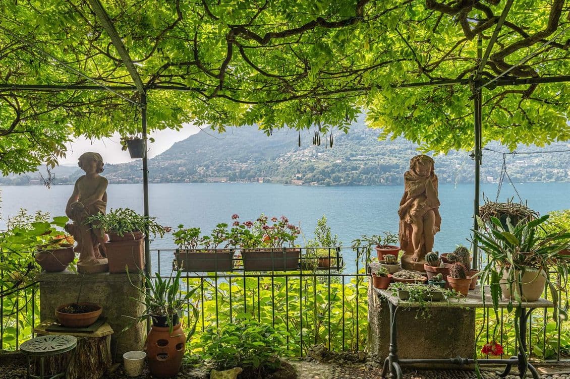 Terrace with a pergola, stone statues and potted plants overlooking Lake Como and the surrounding mountains.