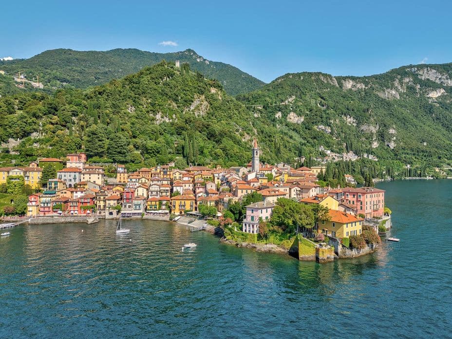 Picturesque village with colourful houses and a church tower on the shore of Lake Como, surrounded by wooded mountains.