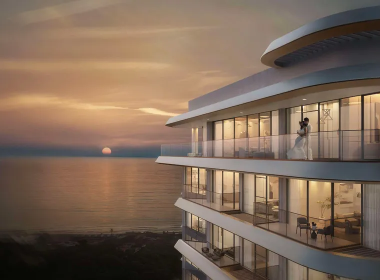 Sunset over the ocean beside a futuristic curved glass building; a bride and groom stand on an illuminated balcony.