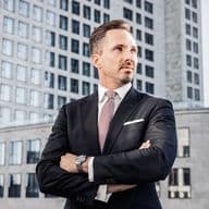 Benjamin Rogmans in a suit stands confidently with arms crossed in front of a cityscape, featuring modern office buildings and a clear sky.