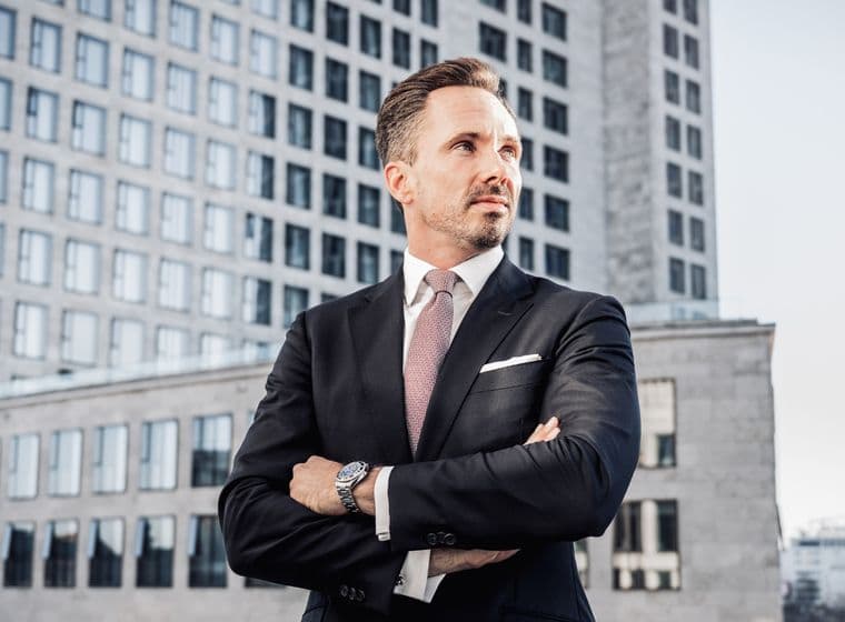 Benjamin Rogmans in a suit stands confidently with arms crossed in front of a cityscape, featuring modern office buildings and a clear sky.
