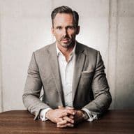 Benjamin Rogmans in a gray suit sits at a wooden table, hands clasped, with a focused expression against a concrete wall background.