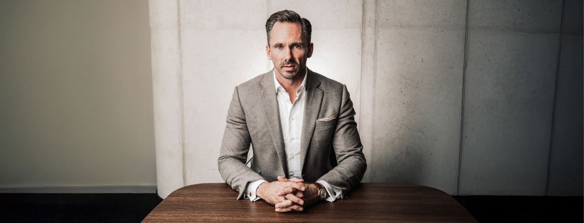 Benjamin Rogmans in a gray suit sits at a wooden table, hands clasped, with a focused expression against a concrete wall background.