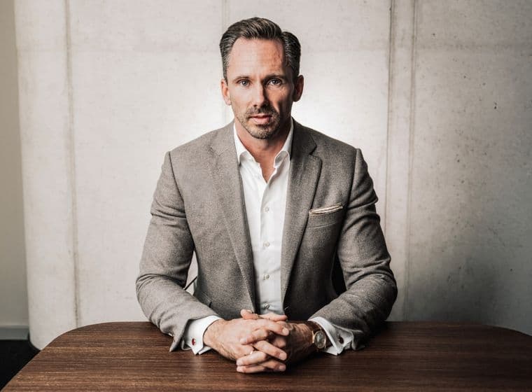 Benjamin Rogmans in a gray suit sits at a wooden table, hands clasped, with a focused expression against a concrete wall background.