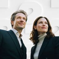 Ein Mann und eine Frau in Business-Kleidung blicken selbstbewusst nach oben, während sie vor einem Hintergrund aus großen weißen Fragezeichen stehen.