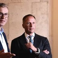 Rackham F. Schröder und Stefan Evers in Anzügen stehen neben einer Steinmauer; der eine mit Brille gestikuliert beim Sprechen, der andere hört mit verschränkten Armen zu.