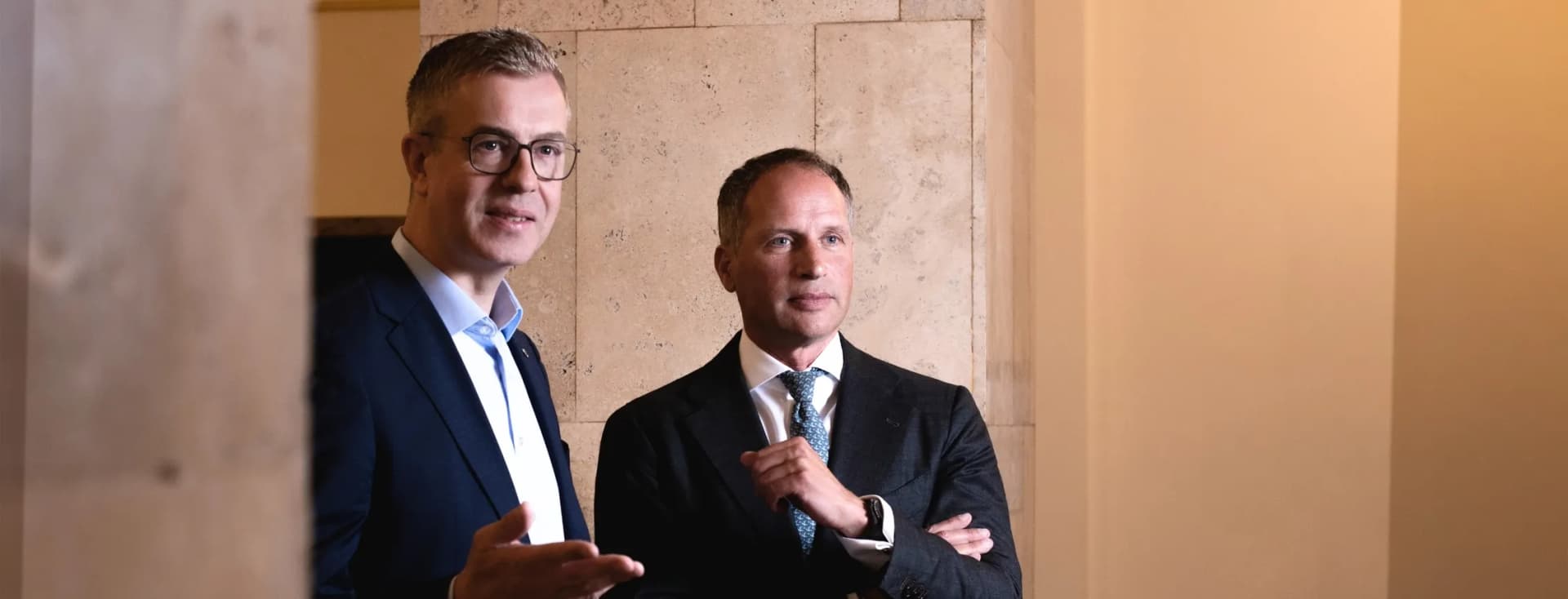 Rackham F. Schröder und Stefan Evers in Anzügen stehen neben einer Steinmauer; der eine mit Brille gestikuliert beim Sprechen, der andere hört mit verschränkten Armen zu.