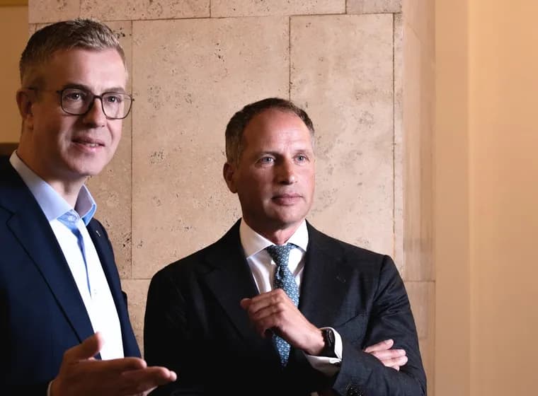 Rackham F. Schröder und Stefan Evers in Anzügen stehen neben einer Steinmauer; der eine mit Brille gestikuliert beim Sprechen, der andere hört mit verschränkten Armen zu.