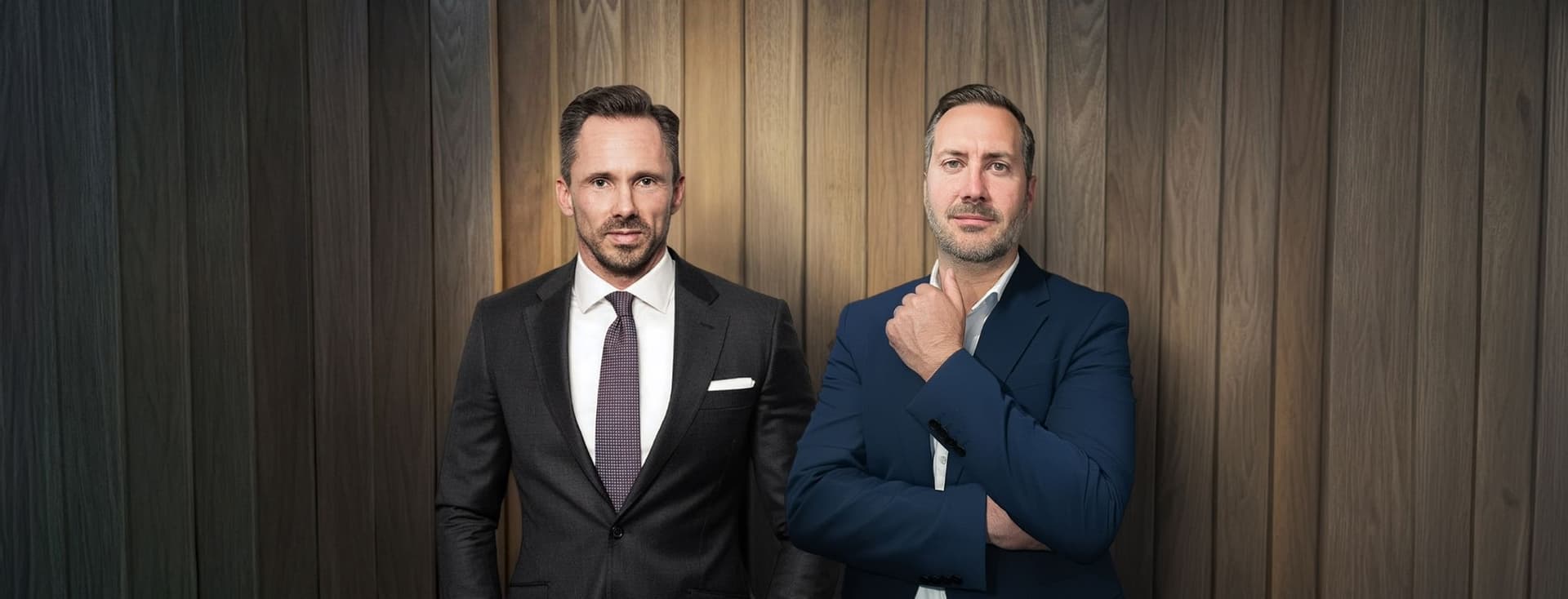 Benjamin Rogmans und Michael Gassmann in eleganten Anzügen stehen selbstbewusst vor einer holzgetäfelten Wand und strahlen eine professionelle und gelassene Haltung aus.