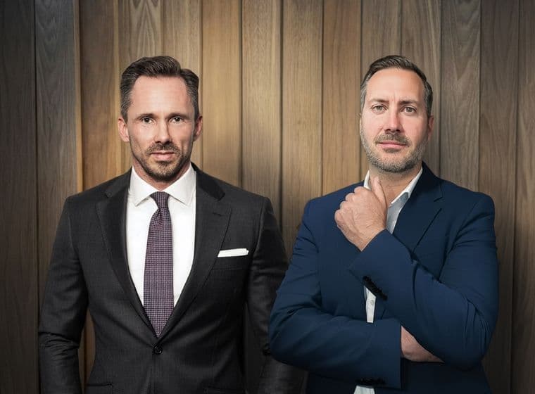 Benjamin Rogmans und Michael Gassmann in eleganten Anzügen stehen selbstbewusst vor einer holzgetäfelten Wand und strahlen eine professionelle und gelassene Haltung aus.