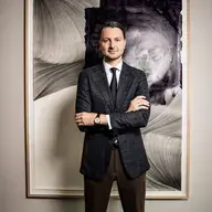 Frederic Weber in einem marineblauen karierten Blazer, einem weißen Hemd und einer schwarzen Krawatte steht mit verschränkten Armen vor einem großen, gerahmten abstrakten Gemälde.