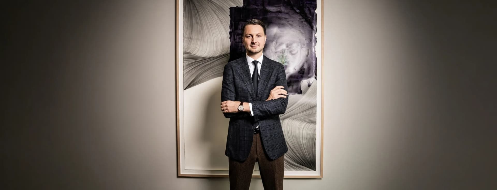 Frederic Weber in einem marineblauen karierten Blazer, einem weißen Hemd und einer schwarzen Krawatte steht mit verschränkten Armen vor einem großen, gerahmten abstrakten Gemälde.