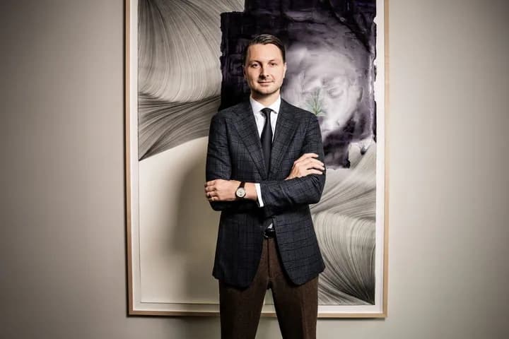 Frederic Weber in einem marineblauen karierten Blazer, einem weißen Hemd und einer schwarzen Krawatte steht mit verschränkten Armen vor einem großen, gerahmten abstrakten Gemälde.