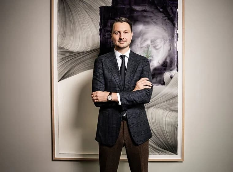 Frederic Weber in einem marineblauen karierten Blazer, einem weißen Hemd und einer schwarzen Krawatte steht mit verschränkten Armen vor einem großen, gerahmten abstrakten Gemälde.