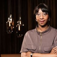 Aissatou Frisch-Baldé mit Brille und kurzen Haaren, die Arme verschränkt, vor einem dunkel gestreiften Hintergrund mit hängenden Pendelleuchten.