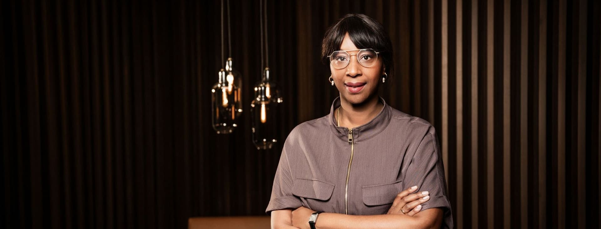 Aissatou Frisch-Baldé mit Brille und kurzen Haaren, die Arme verschränkt, vor einem dunkel gestreiften Hintergrund mit hängenden Pendelleuchten.