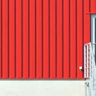 Rotes Wellblechgebäude mit weißer Tür und Treppe, angrenzend an einen kleinen Abschnitt einer grauen Wand auf der linken Seite.