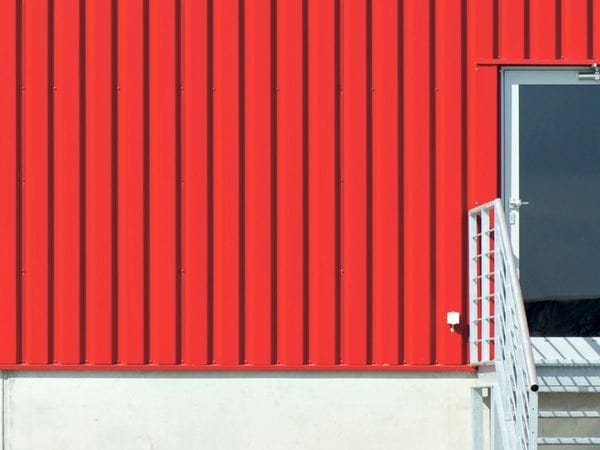 Rotes Wellblechgebäude mit weißer Tür und Treppe, angrenzend an einen kleinen Abschnitt einer grauen Wand auf der linken Seite.