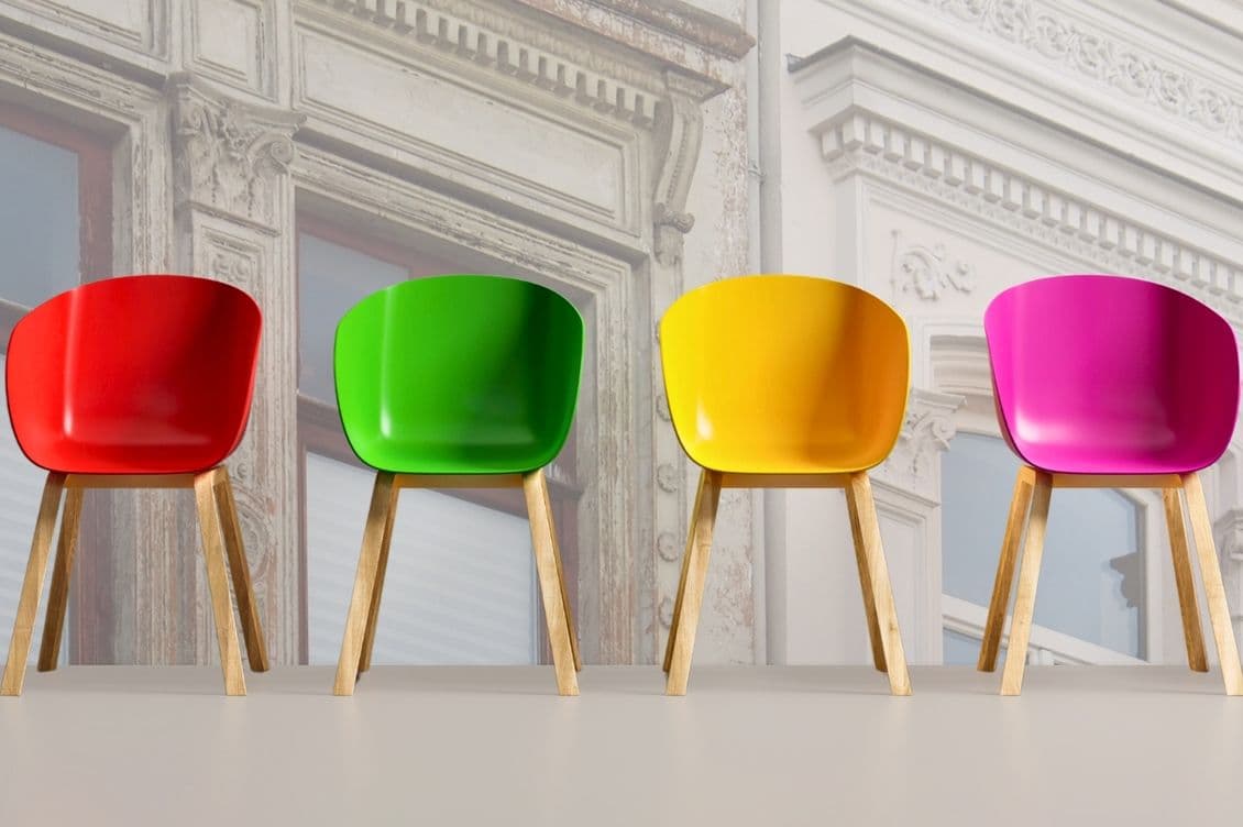 Sechs bunte Stühle mit Holzbeinen standen auf einem hellen Boden in einer Reihenfolge, die den Farben des Regenbogens von Schwarz bis Blau folgte, vor dem Hintergrund einer verblassten, verzierten Gebäudefassade.