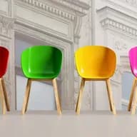 Sechs bunte Stühle mit Holzbeinen standen auf einem hellen Boden in einer Reihenfolge, die den Farben des Regenbogens von Schwarz bis Blau folgte, vor dem Hintergrund einer verblassten, verzierten Gebäudefassade.