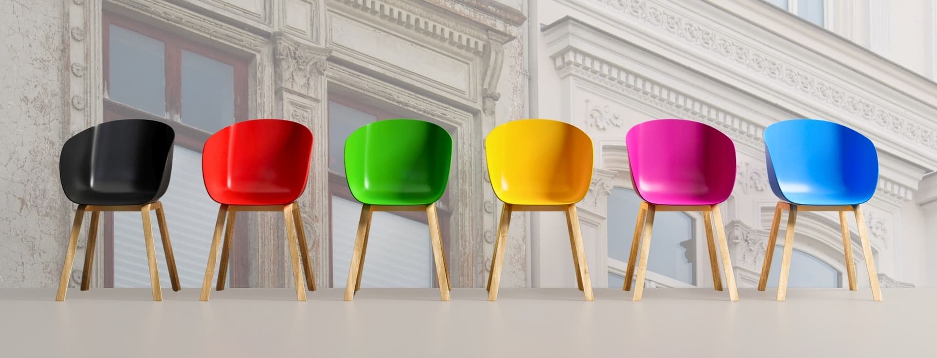 Sechs bunte Stühle mit Holzbeinen standen auf einem hellen Boden in einer Reihenfolge, die den Farben des Regenbogens von Schwarz bis Blau folgte, vor dem Hintergrund einer verblassten, verzierten Gebäudefassade.
