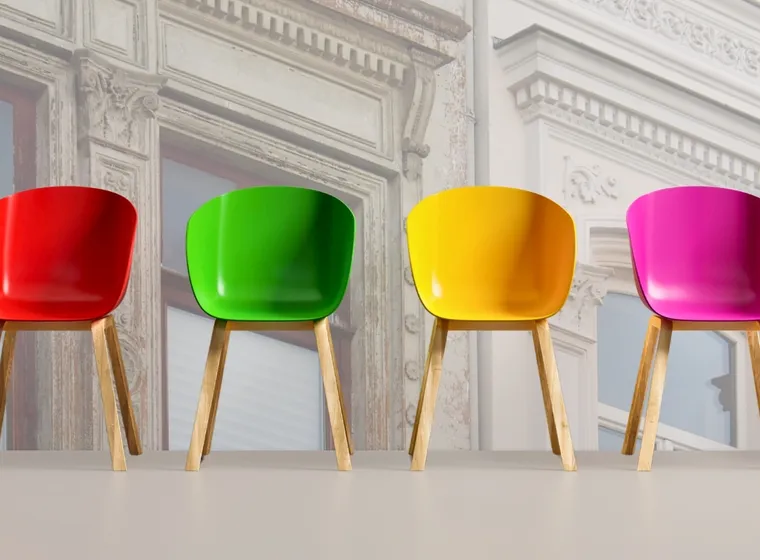 Sechs bunte Stühle mit Holzbeinen standen auf einem hellen Boden in einer Reihenfolge, die den Farben des Regenbogens von Schwarz bis Blau folgte, vor dem Hintergrund einer verblassten, verzierten Gebäudefassade.