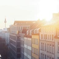 Sunlit cityscape with residential buildings, balconies, and a visible distant TV tower under a clear sky, creating a warm, serene atmosphere.