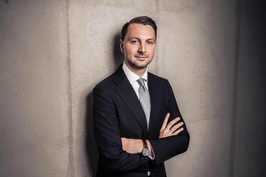 Man in a suit with arms crossed, standing against a concrete wall, wearing a light blue tie and watch, gazing confidently at the camera.