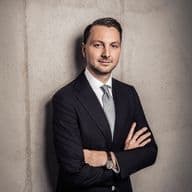Man in a suit with arms crossed, standing against a concrete wall, wearing a light blue tie and watch, gazing confidently at the camera.