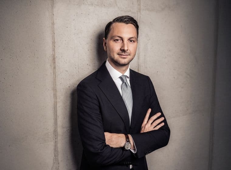 Man in a suit with arms crossed, standing against a concrete wall, wearing a light blue tie and watch, gazing confidently at the camera.