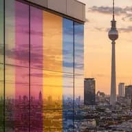 Berliner Stadtansicht bei Sonnenuntergang mit einem Glasgebäude aus vertikal angeordneten Regenbogenscheiben; die Berliner Skyline wird vom Fernsehturm und dem Berliner Dom geprägt.