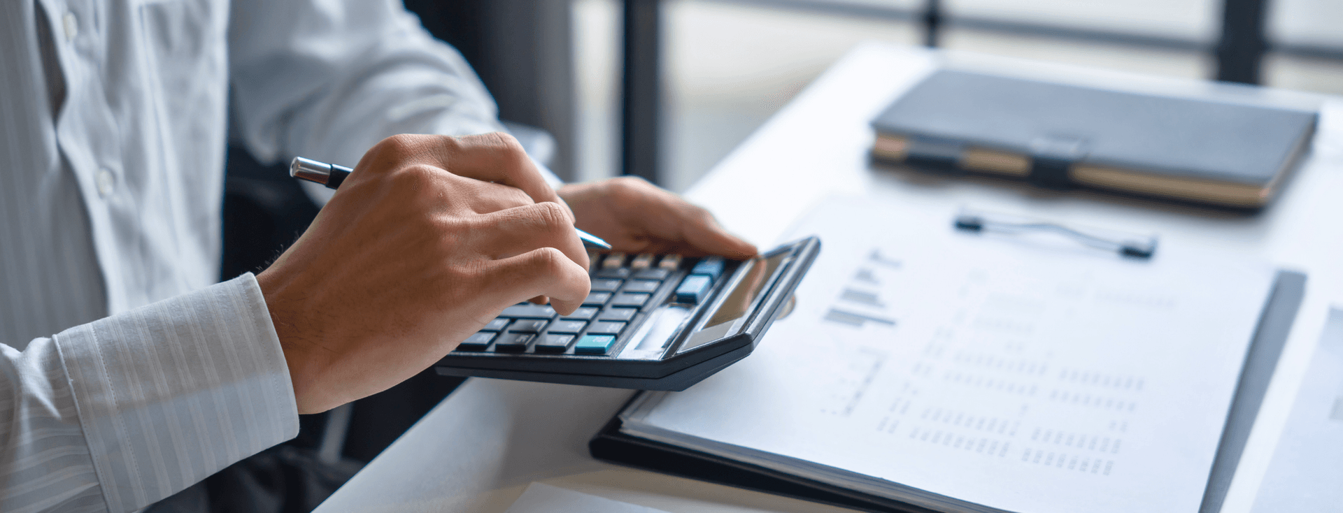 A person in a white shirt uses a calculator and holds a pen, with documents and a notebook on the desk.
