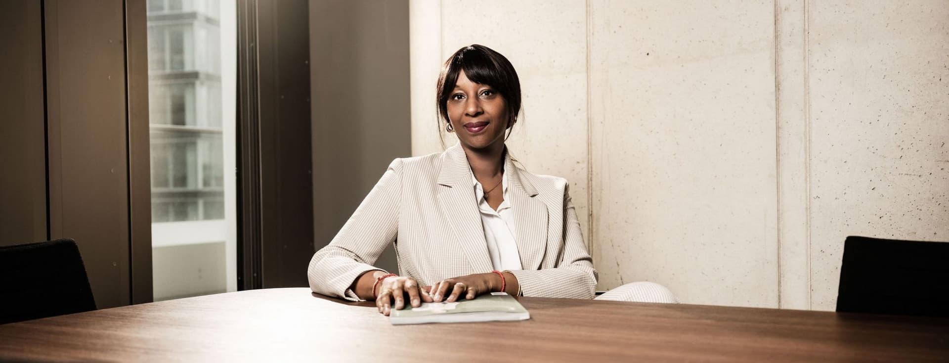 Aissatou Frisch-Baldé sitzt in einem hellen Kostüm mit einem geschlossenen Buch an einem Tisch vor einer modernen Kulisse.