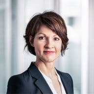 Dr. Andrea Peters mit kurzen braunen Haaren in einem schwarzen Blazer und einer weißen Bluse vor einem unscharfen, hellen Hintergrund.