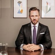 Benjamin Rogmans sitzt im Anzug an einem Holztisch, hinter ihm hängt ein abstraktes Gemälde an der Wand. Auf dem Tisch steht eine Glaskaraffe.