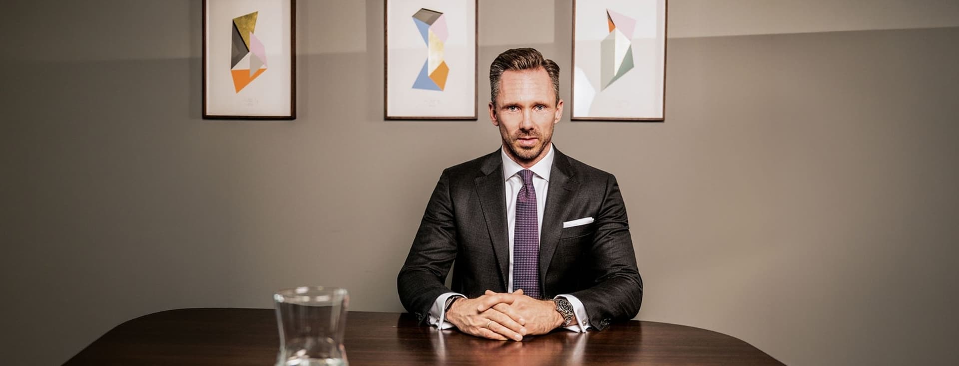 Benjamin Rogmans sitzt im Anzug an einem Holztisch, hinter ihm hängt ein abstraktes Gemälde an der Wand. Auf dem Tisch steht eine Glaskaraffe.