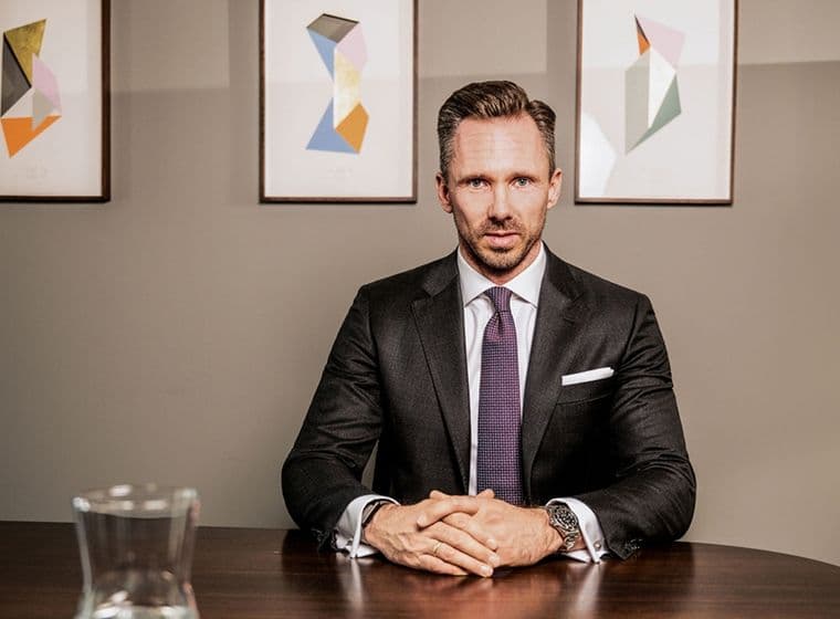 Benjamin Rogmans sitzt im Anzug an einem Holztisch, hinter ihm hängt ein abstraktes Gemälde an der Wand. Auf dem Tisch steht eine Glaskaraffe.