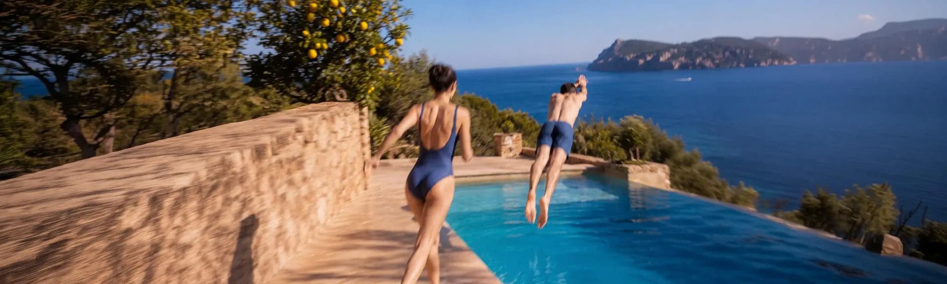 A couple dives into an ocean-view infinity pool from the stone terrace of a luxury villa, shaded by a lemon tree