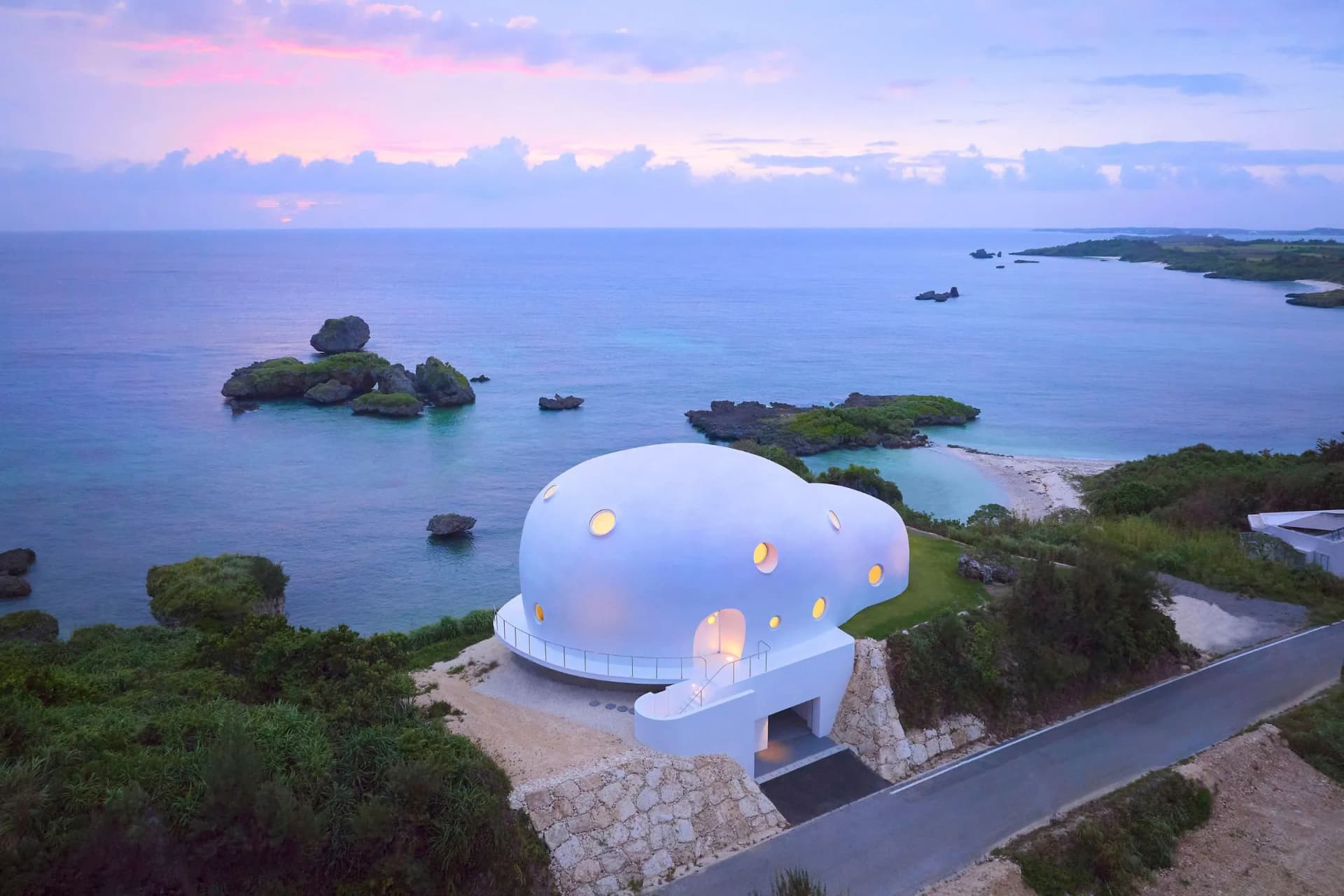 White domed house with rounded windows sits on a rocky coastal shore, overlooking a calm blue sea at sunset, with a winding road nearby.
