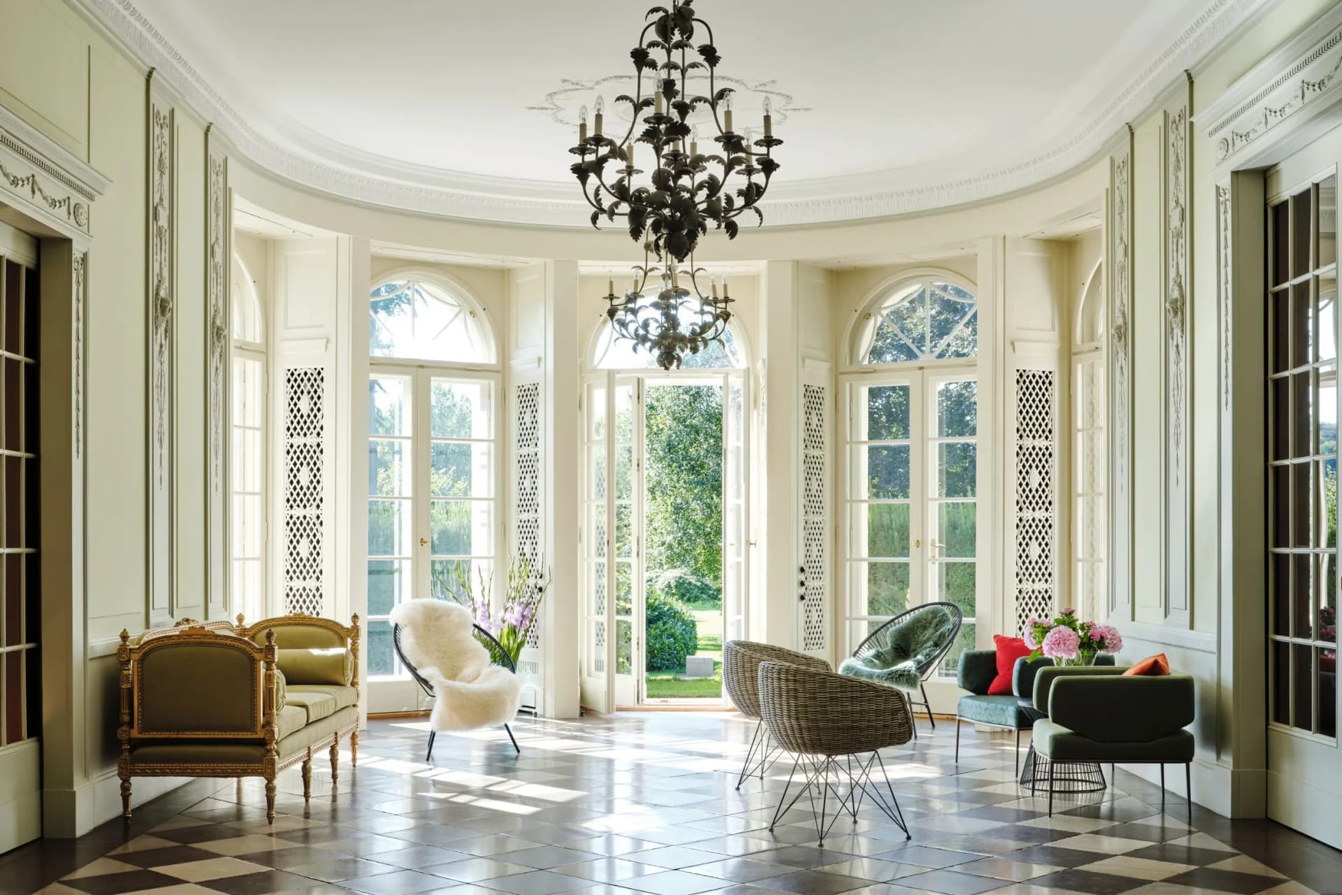An elegant, sun-drenched drawing room with ornate mouldings, a large wrought-iron chandelier, arched windows and wicker chairs; chequered floor.