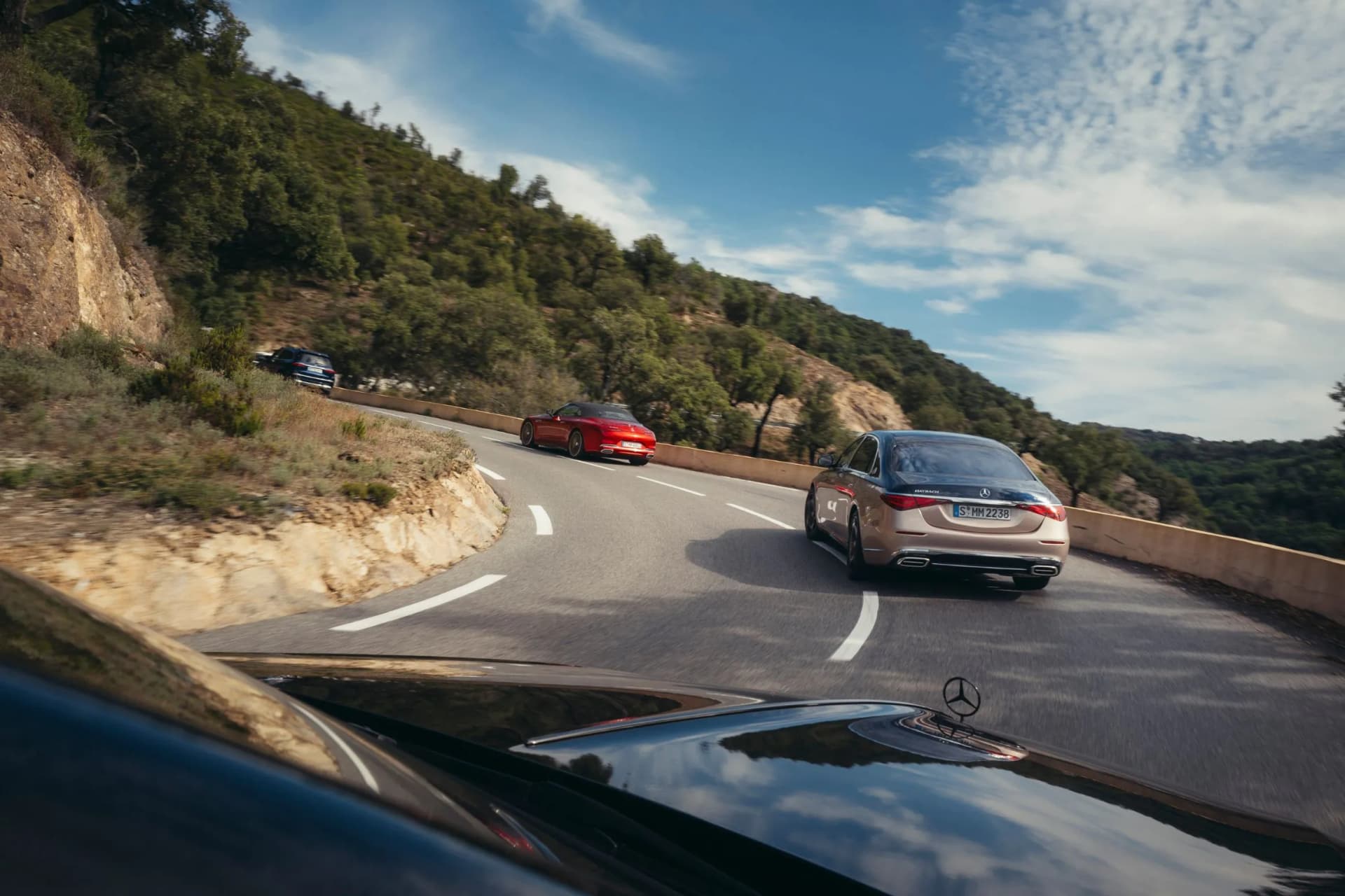 Several cars are driving along a winding mountain road; to the left is a rocky slope, to the right a crash barrier, and above them the blue sky.