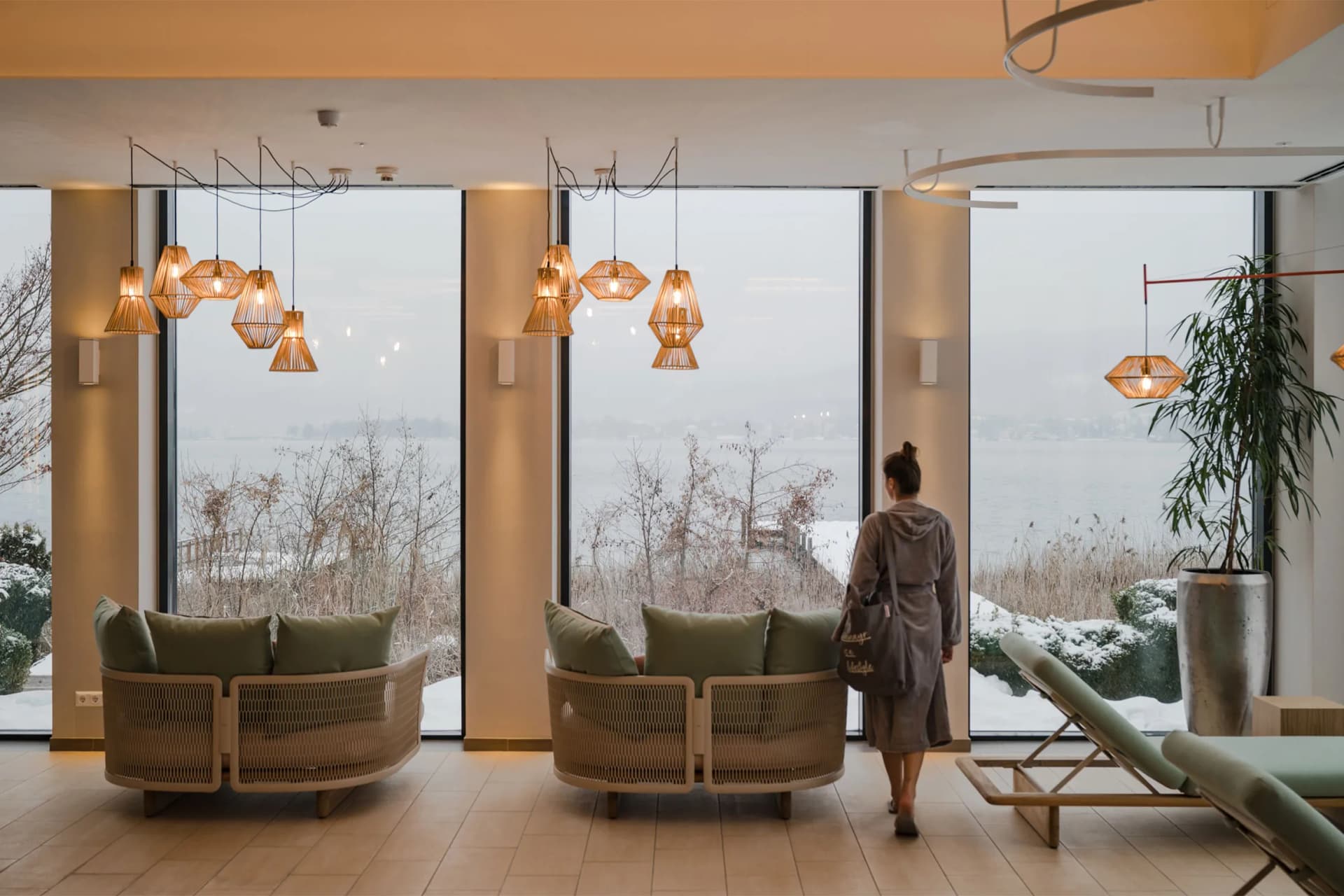 Cozy modern lounge with hanging geometric lamps and circular wicker sofas; a person in a hoodie stands by large windows overlooking a snowy landscape.